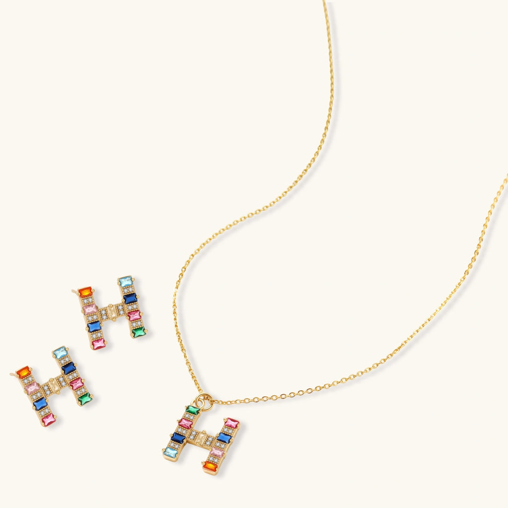 Gold-plated H initial pendant necklace and stud earrings set with multicolor baguette zirconia stones on cream background.